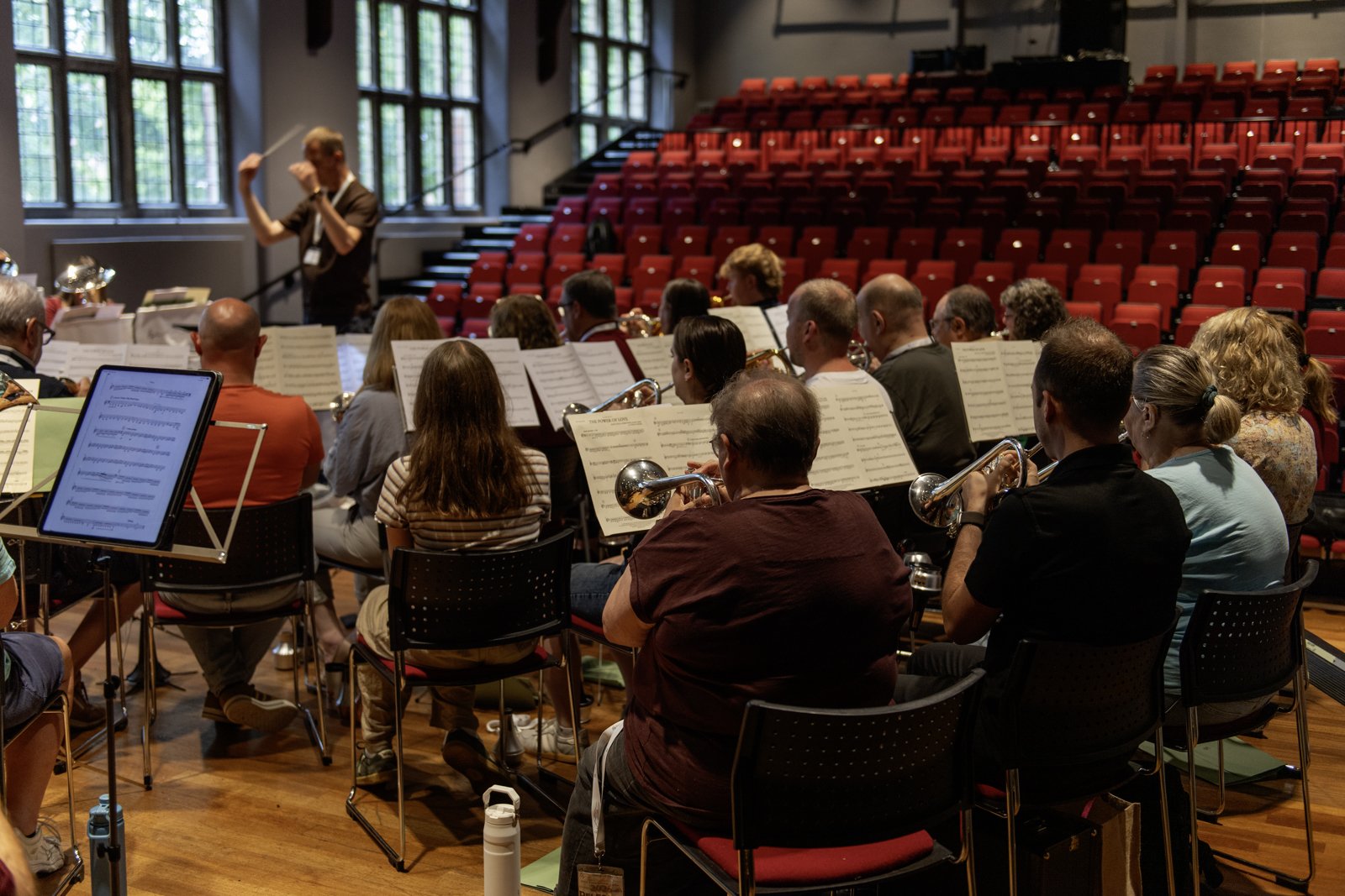 Orchestra rehearsing in a concert hall with red seats and large windows