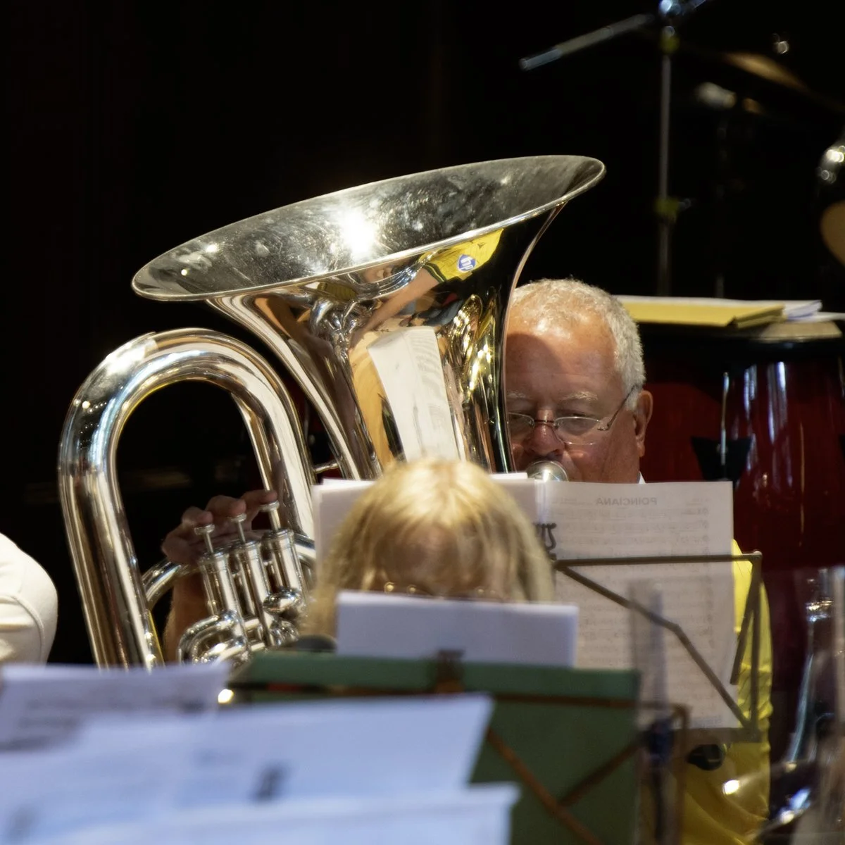 A person playing a silver tuba, with sheet music in front of them, in a dark setting.