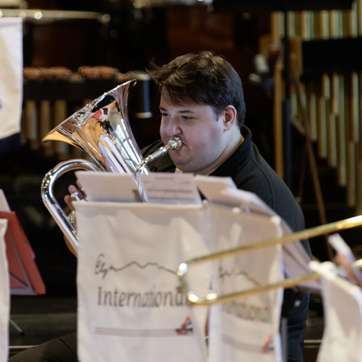 A young man playing a brass instrument, likely a euphonium, during a musical rehearsal or performance in an indoor setting.