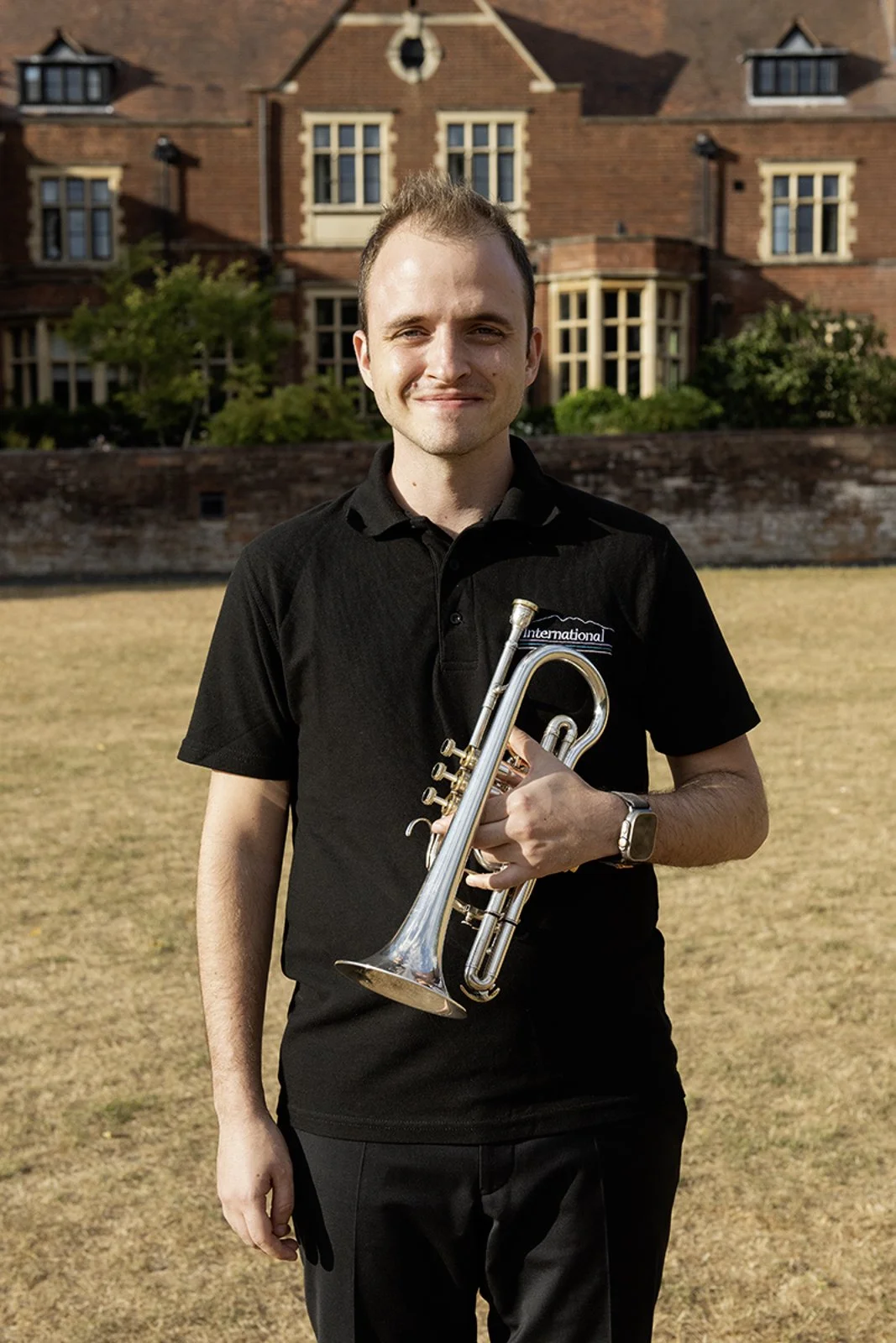 A young man smiling outdoors holding a silver trumpet, standing on a grassy area in front of a brick building with multiple windows and trees.