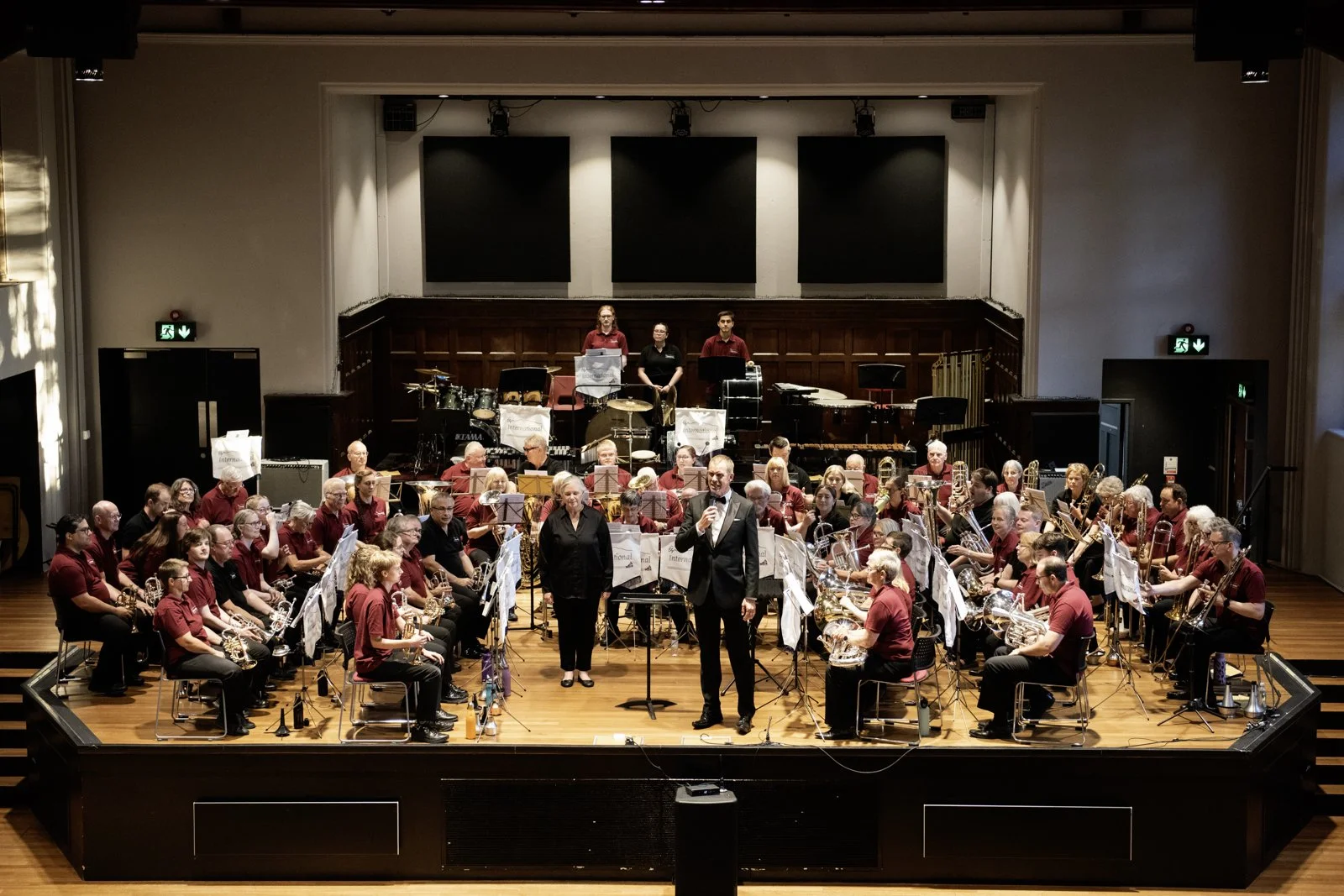 Orchestra performing on stage with conductor holding microphone, surrounded by musicians playing various instruments, in a concert hall.
