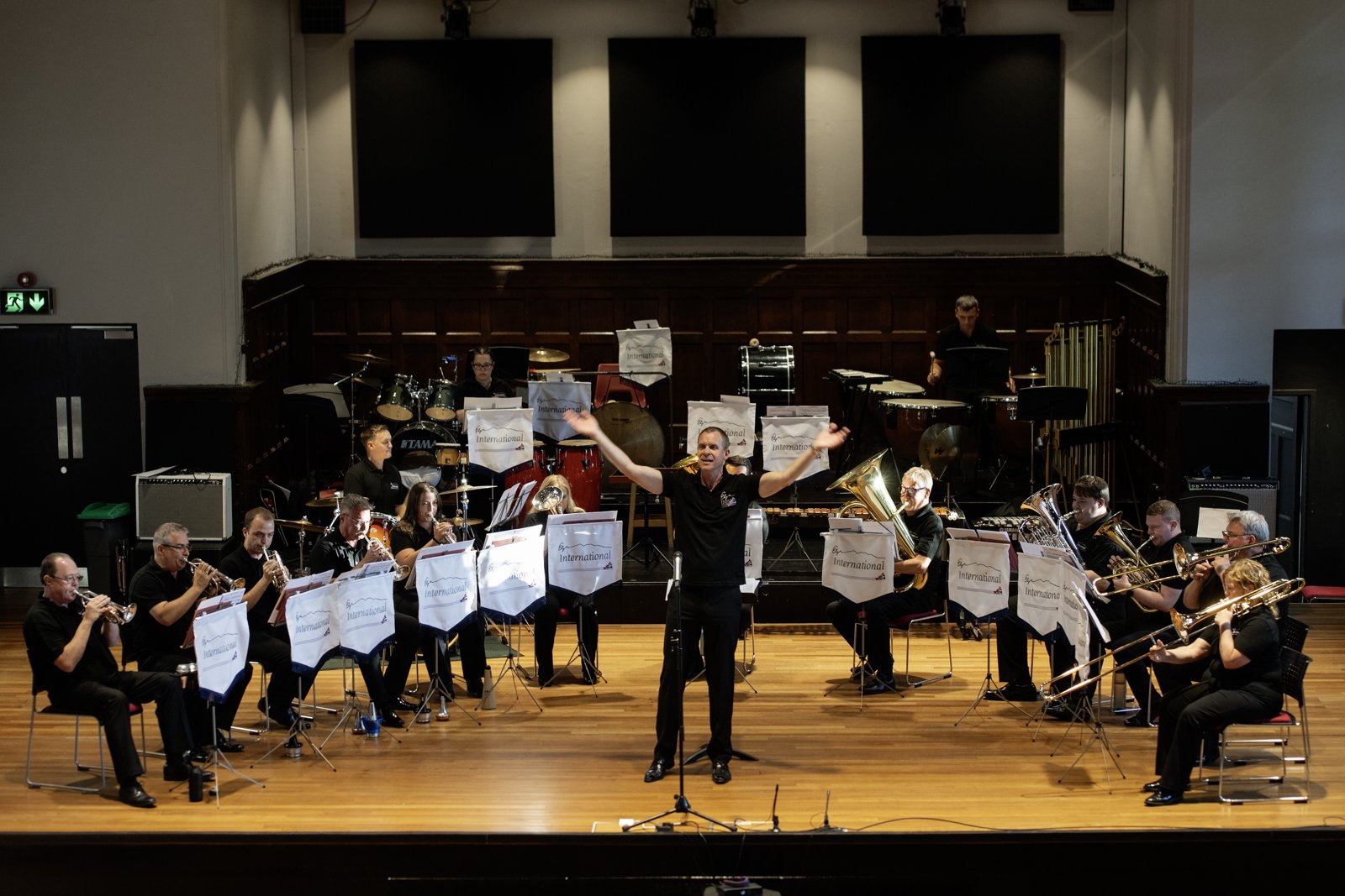 An orchestra performing on stage with a conductor standing in front, raising his arms. The musicians are playing various brass and percussion instruments, and there's a percussion section in the background. The stage has a wooden floor and music stan