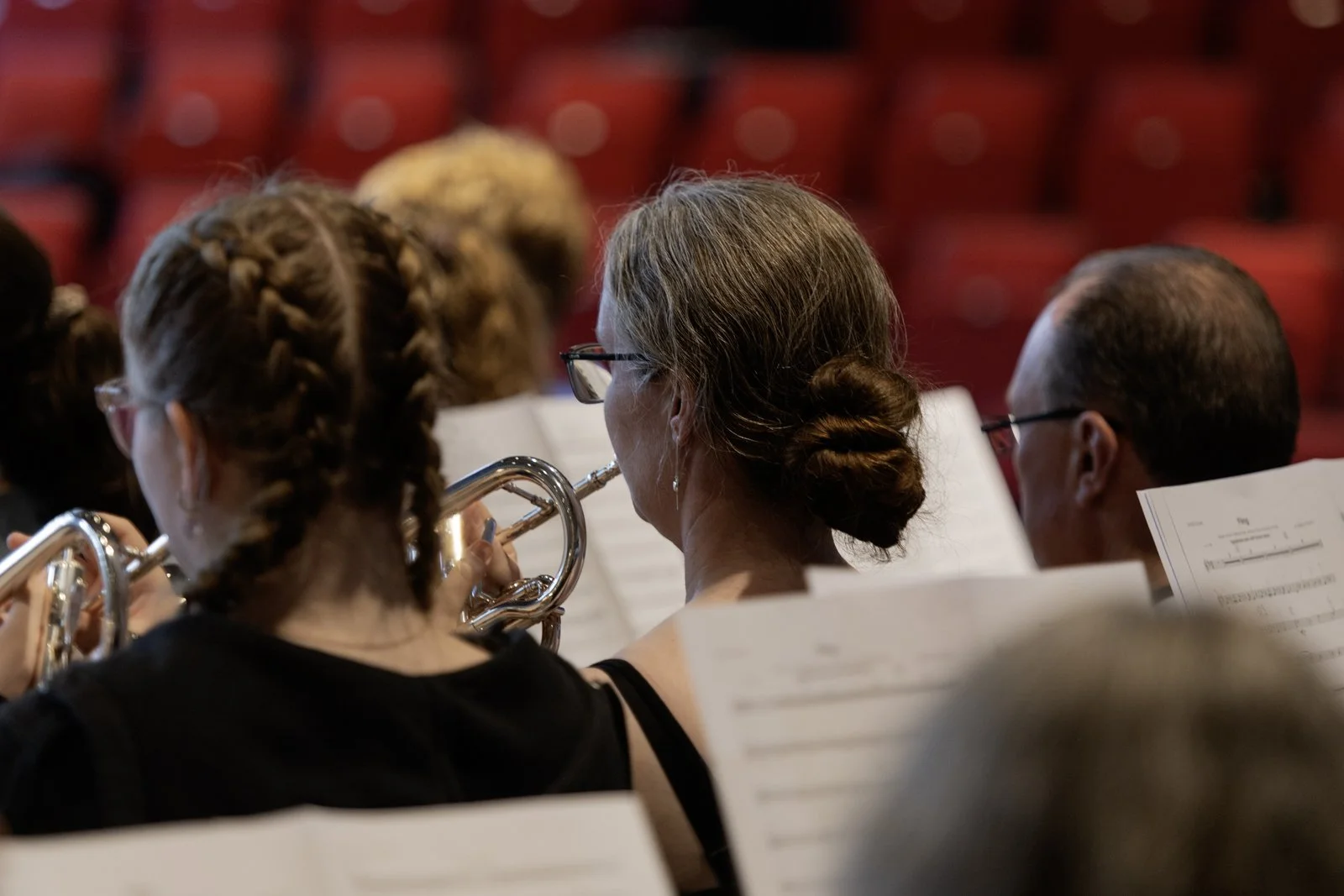 Orchestra members, including a woman with glasses and her hair in a bun, playing wind instruments during a performance, with sheet music and red theater seats in the background.
