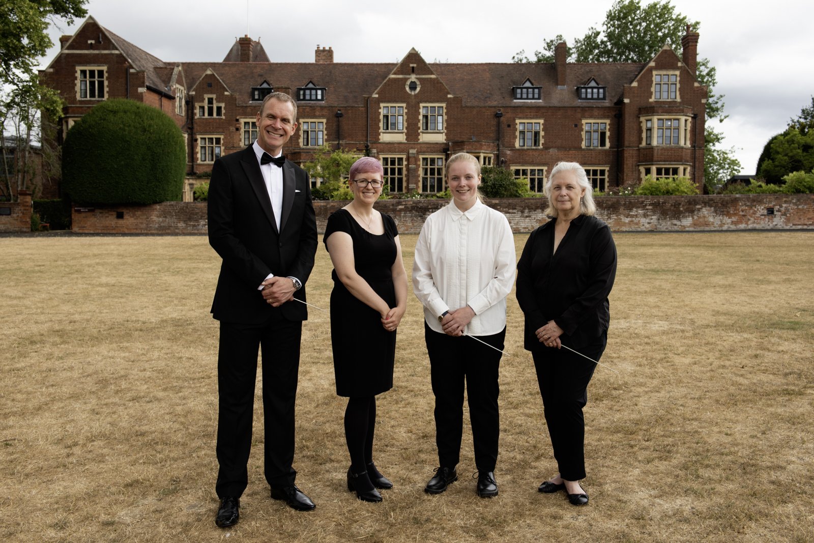 Four people stand outdoors on a dry grassy field in front of a large brick house. They are dressed formally and smiling.