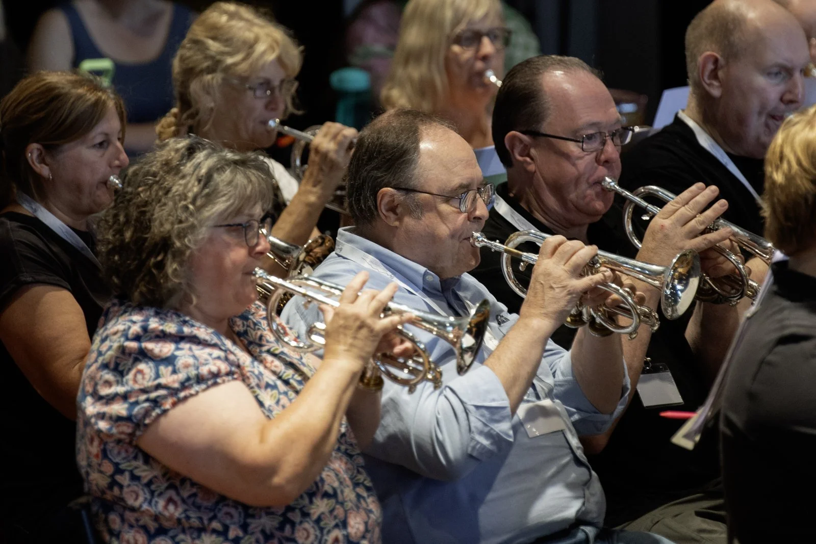 Group of people, mostly middle-aged, sitting and playing small silver trumpets during a musical performance or practice session, with some women visible in the background.