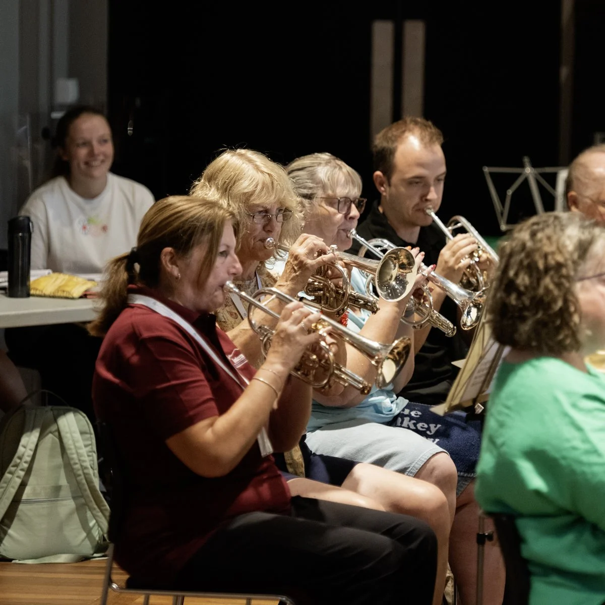 Group of musicians playing trumpets together in a room, with a woman in a red shirt in the foreground and others behind her, some reading sheet music.