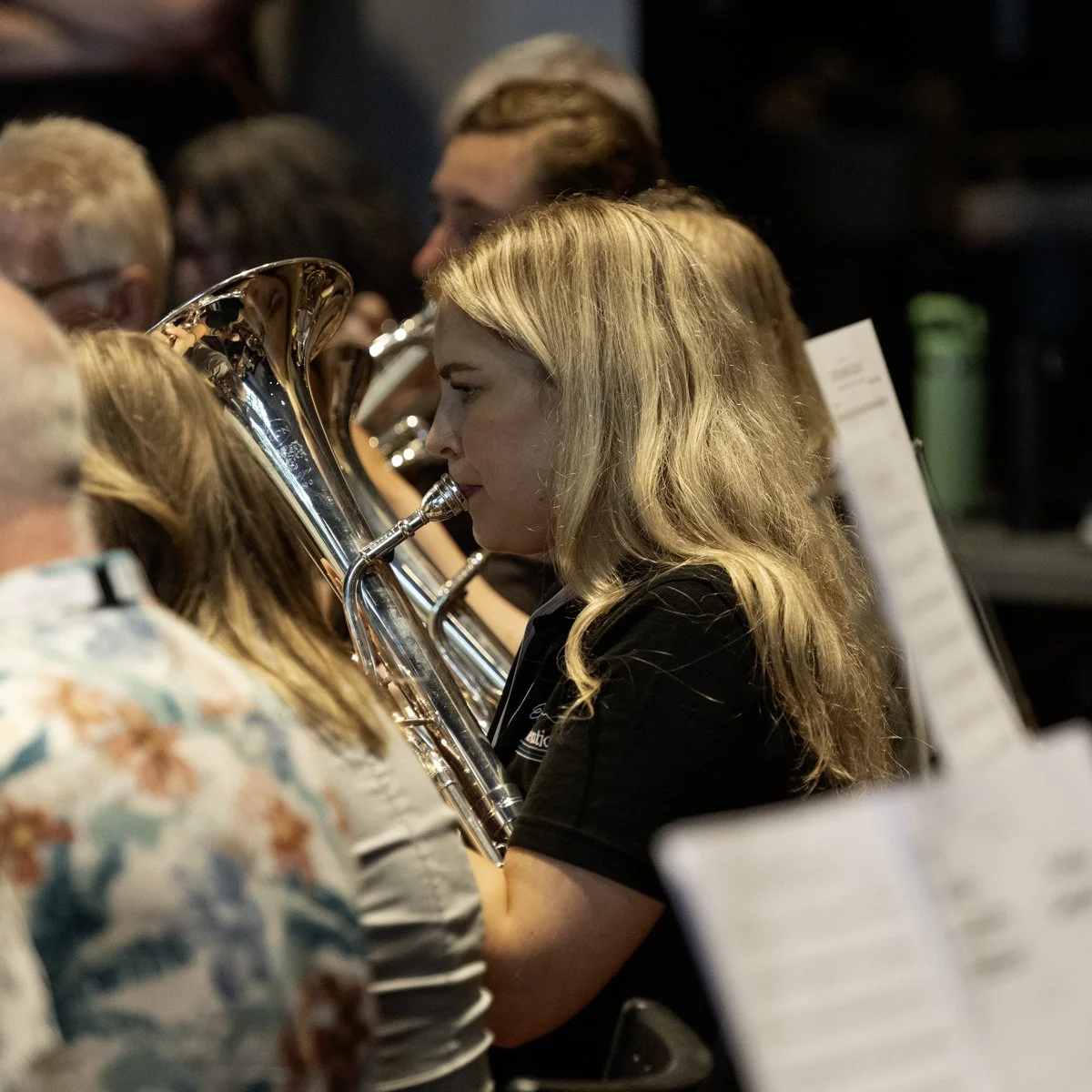A young woman in a black shirt plays a brass instrument in an orchestra, surrounded by other musicians.