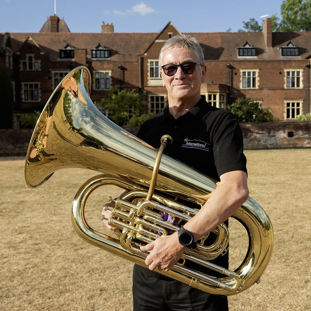 A man wearing sunglasses and a black polo shirt is holding a shiny brass tuba outdoors in front of a large brick building with multiple windows and trees. The sky is clear and blue.