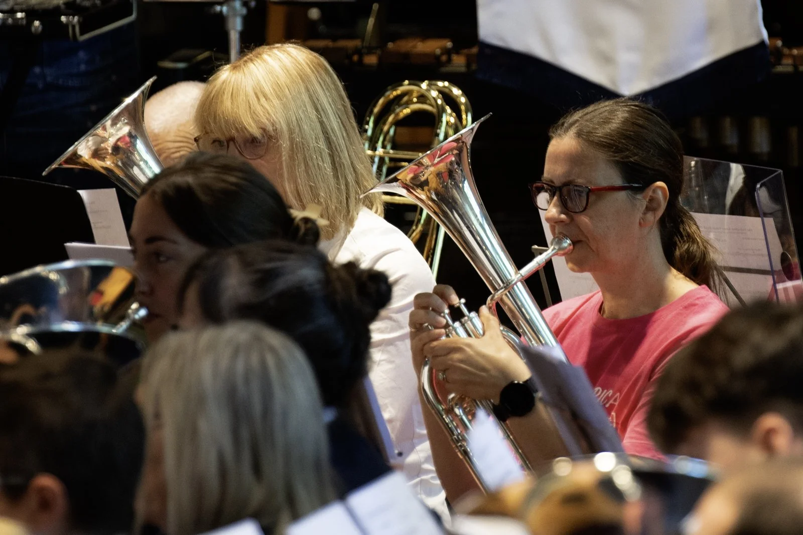 A group of musicians playing brass instruments in an orchestra, with one woman in a pink shirt and glasses prominently featured playing a euphonium or tuba.
