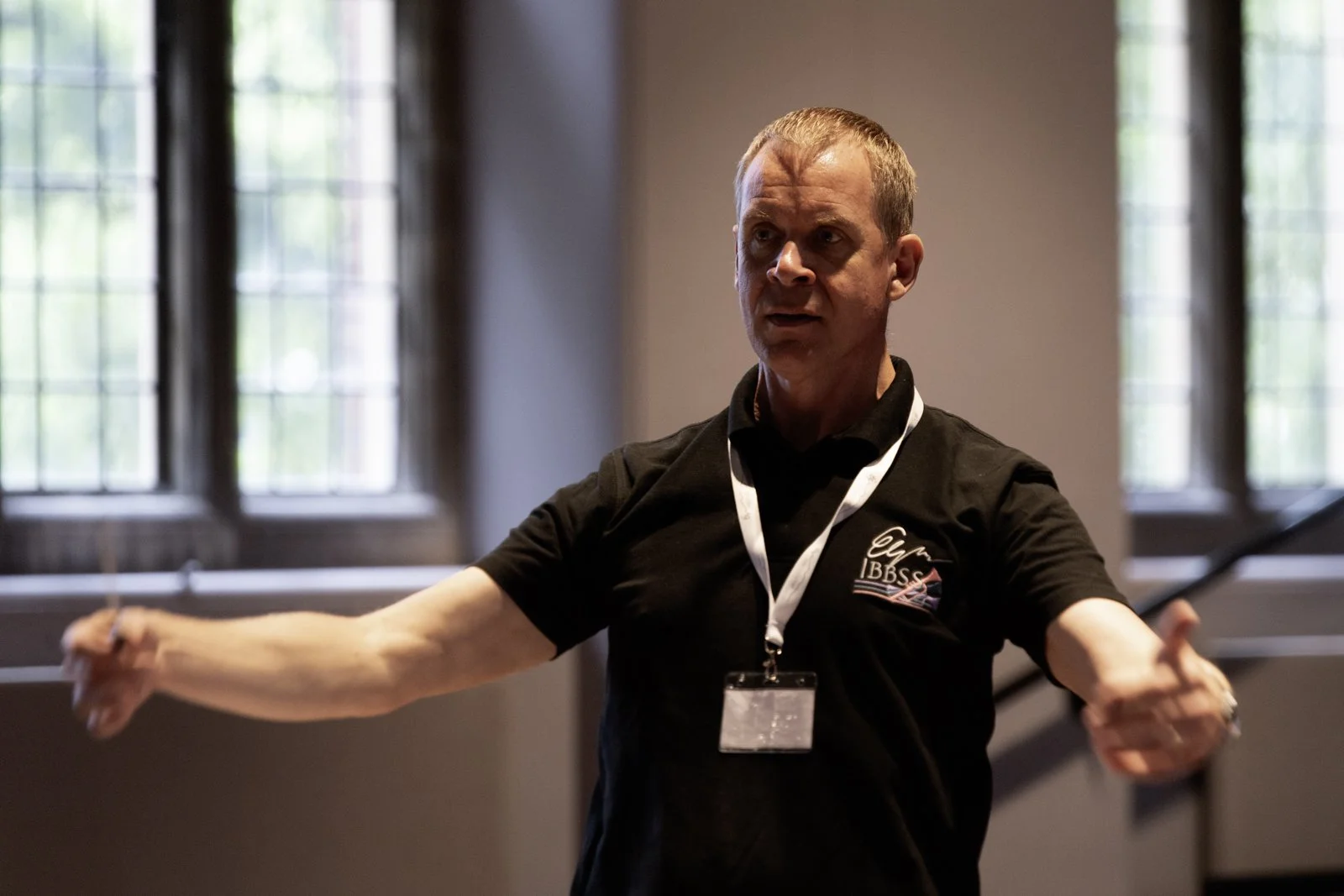 A man with short gray hair wearing a black polo shirt with a logo, gestures with his arms during a presentation or demonstration inside a room with large windows in the background.
