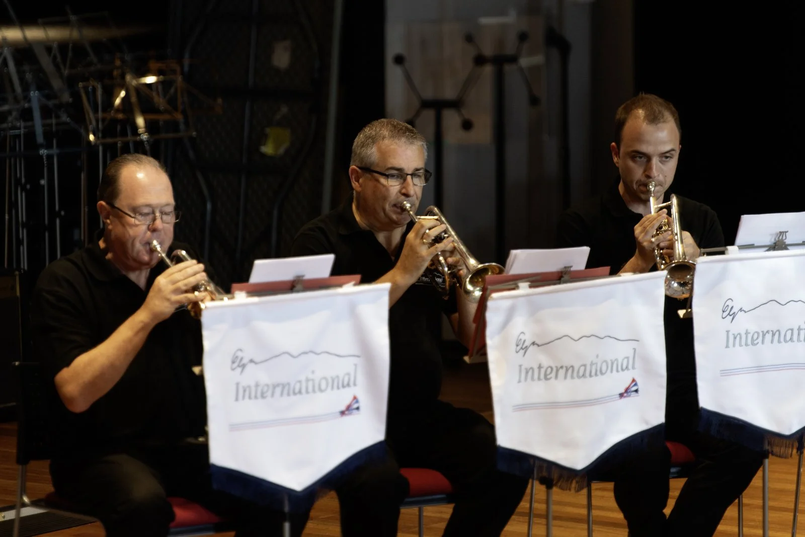 Three men sitting in a row, playing brass instruments during a music performance. They are reading sheet music in front of them with music stands that have 'International' written on them.