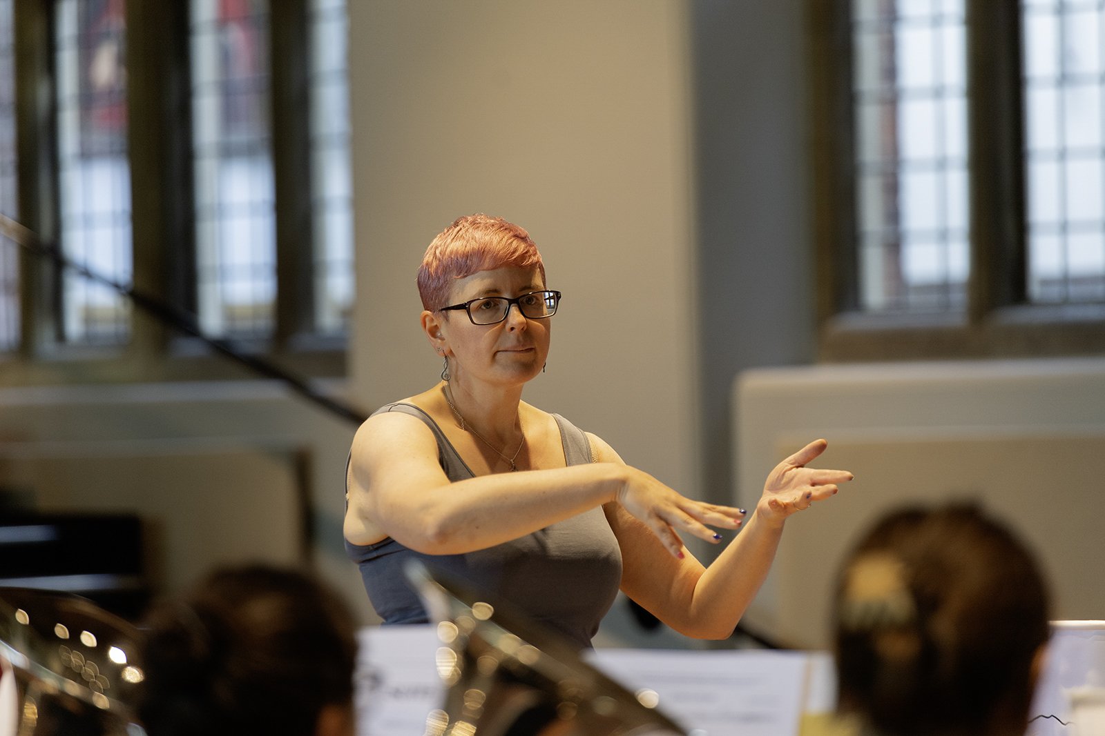 A woman with short pink hair and glasses conducting an orchestra in a room with large windows.