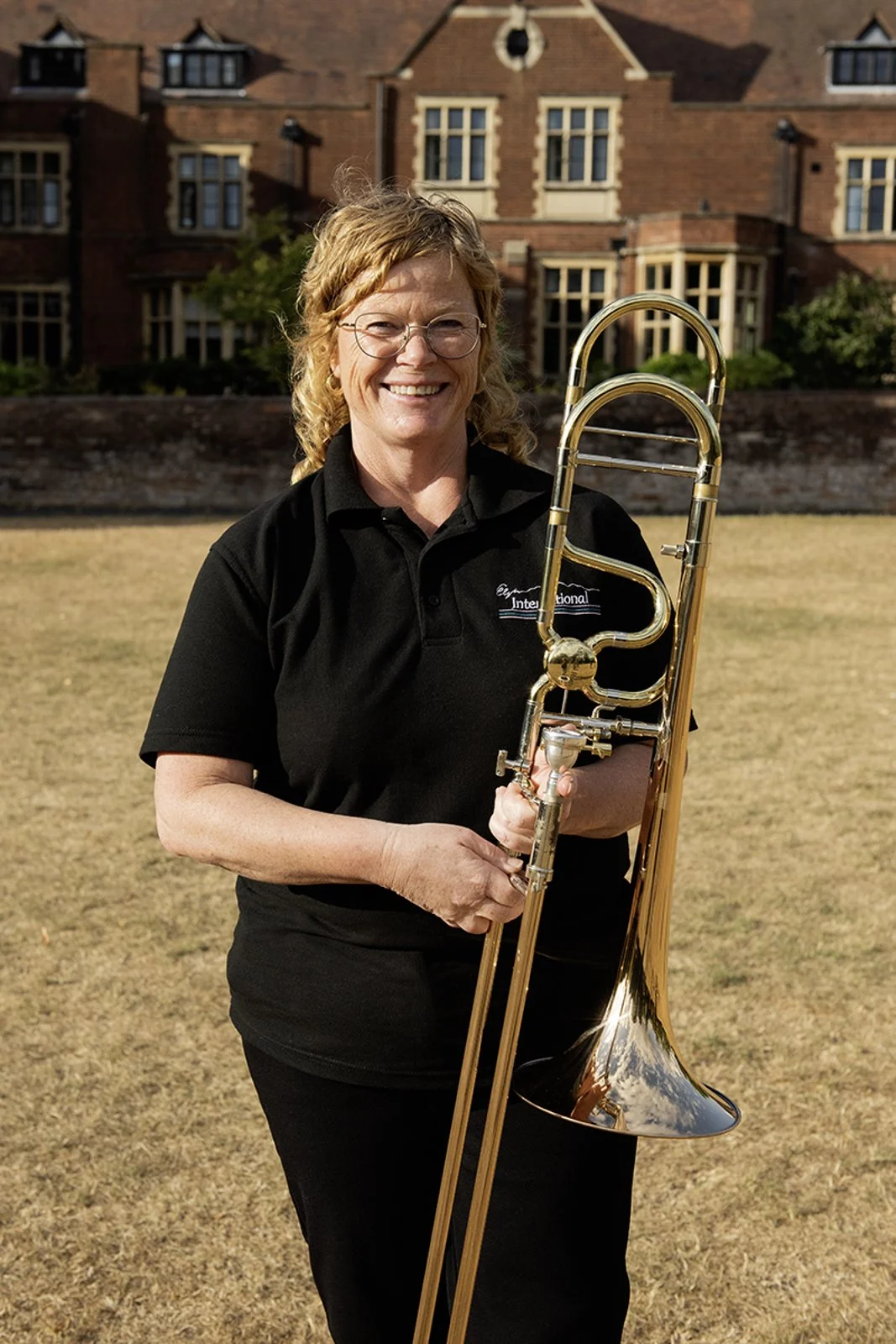 A smiling woman with glasses and curly red hair holding a trombone while standing on a grassy field in front of a large brick building.