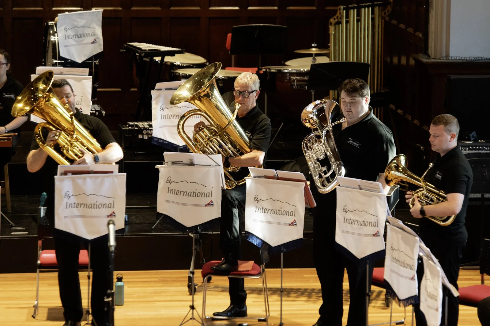 A group of musicians performing on stage, playing brass instruments such as tubas and euphoniums, with music stands marked 'International' in front of them, in a wood-paneled concert hall.
