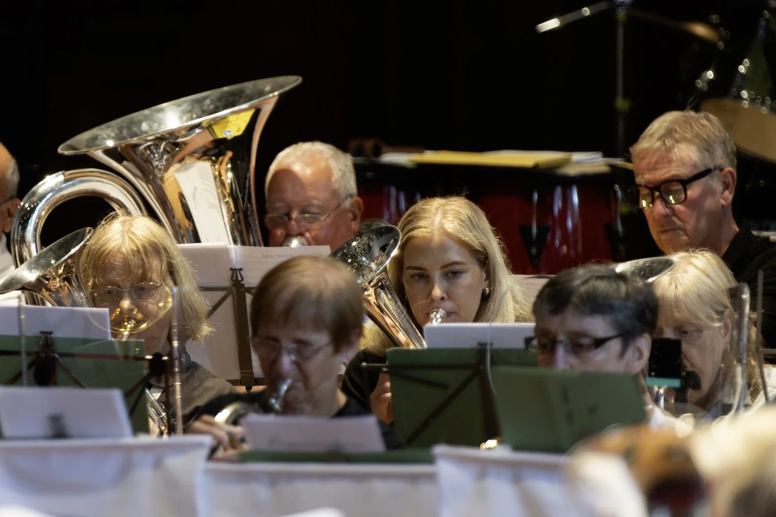 A group of musicians performing in an orchestra, including several women and men playing wind instruments like flutes, clarinets, and horns, with a drum set visible in the background.