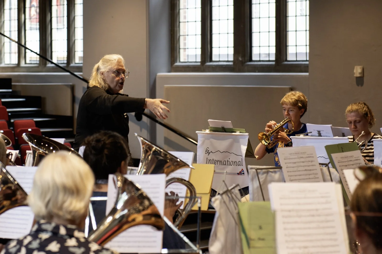 A music conductor leading a group of young musicians in a rehearsal, with a windowed wall letting in natural light.