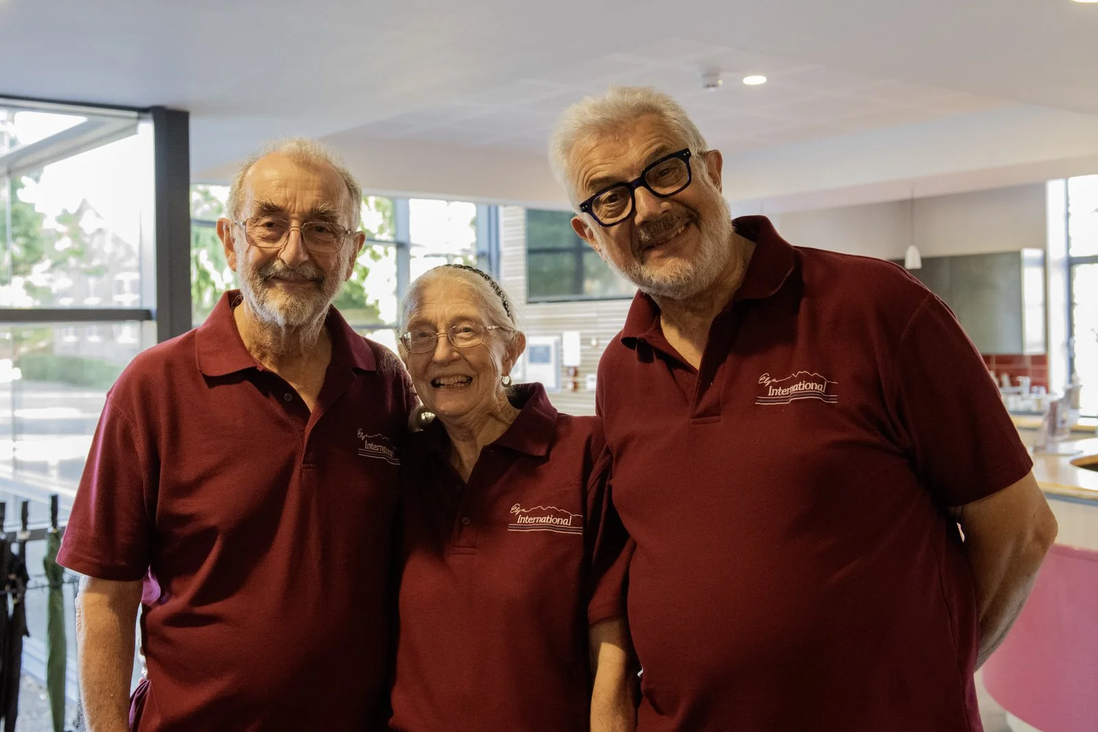 Three elderly people, two men and one woman, smiling and wearing matching maroon polo shirts, standing inside a well-lit room with large windows.