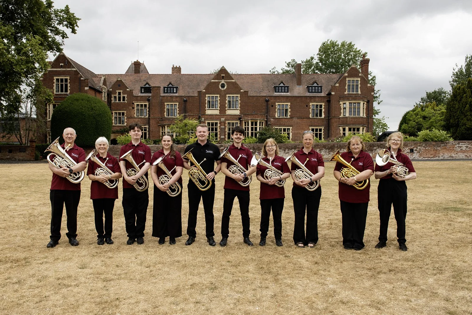 A group of ten people standing outdoors in front of a large brick building. They are dressed in maroon and black shirts, holding brass musical instruments, and smiling at the camera.