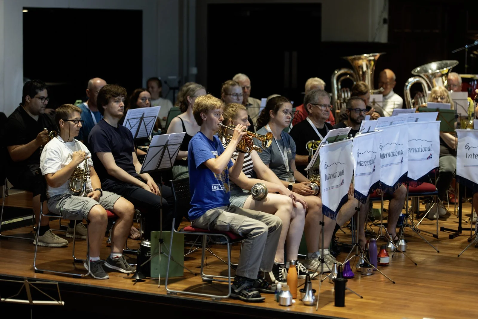 An orchestra rehearsal or performance with musicians of various ages playing brass and wind instruments, seated behind music stands with banners reading 'International.'