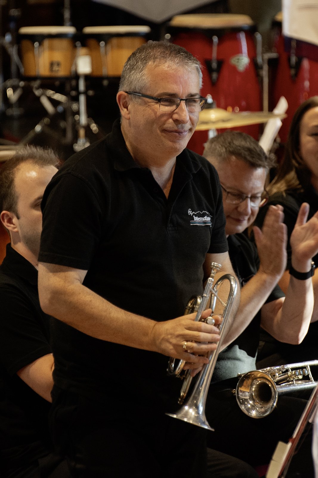 A man holding a trumpet in a music band, surrounded by other musicians clapping, with percussion instruments in the background.