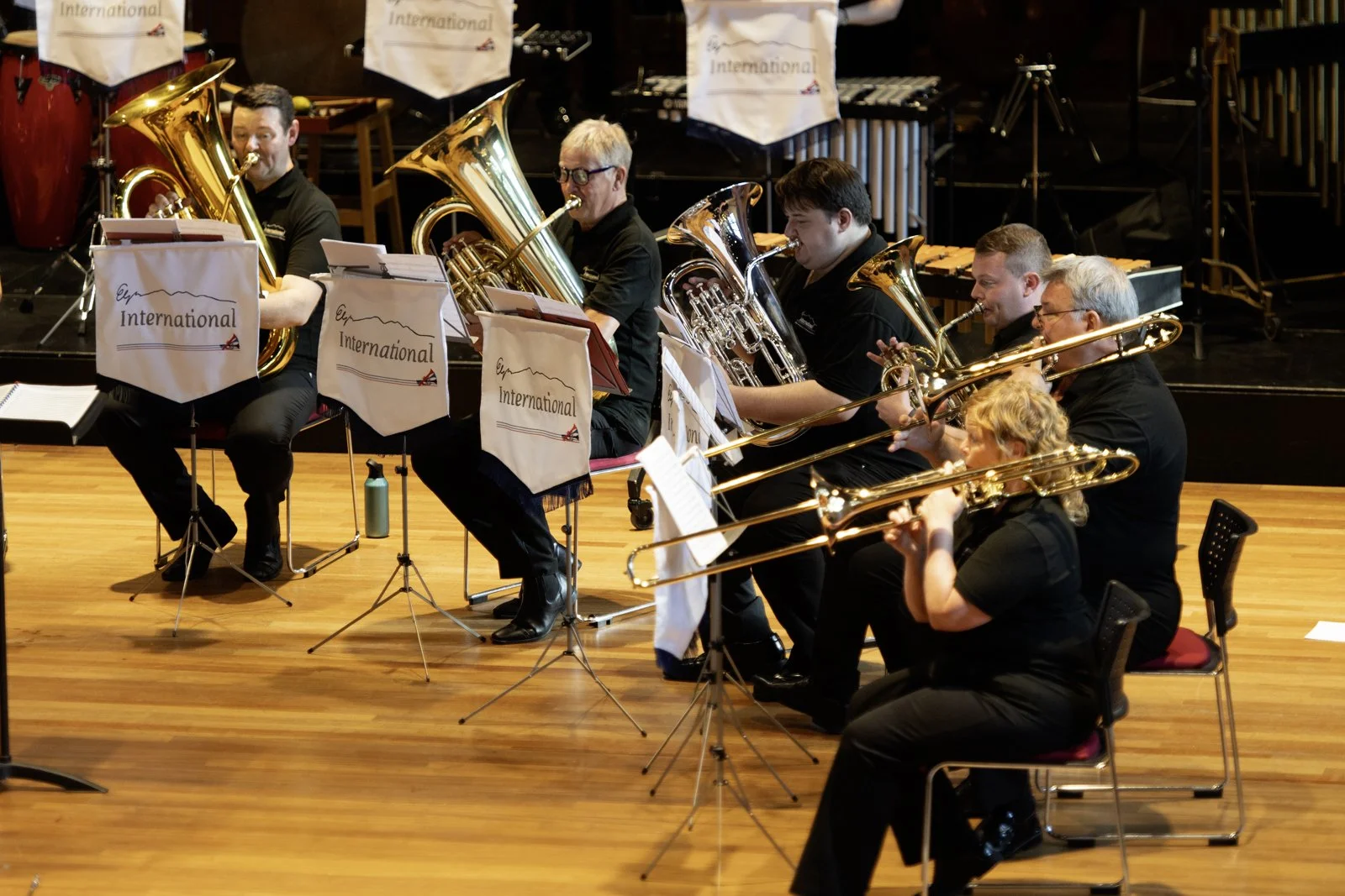 A group of musicians playing brass instruments on a stage. The musicians are seated, reading sheet music, and performing in a concert hall with wooden flooring and curtain in the background.