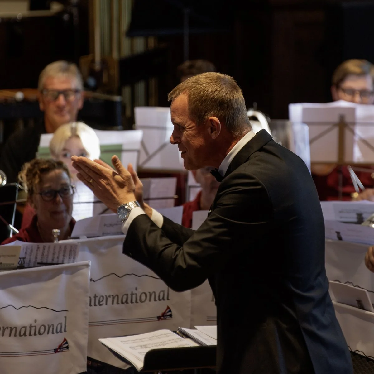 A conductor in a black tuxedo leading an orchestra, with musicians in the background, during a performance.