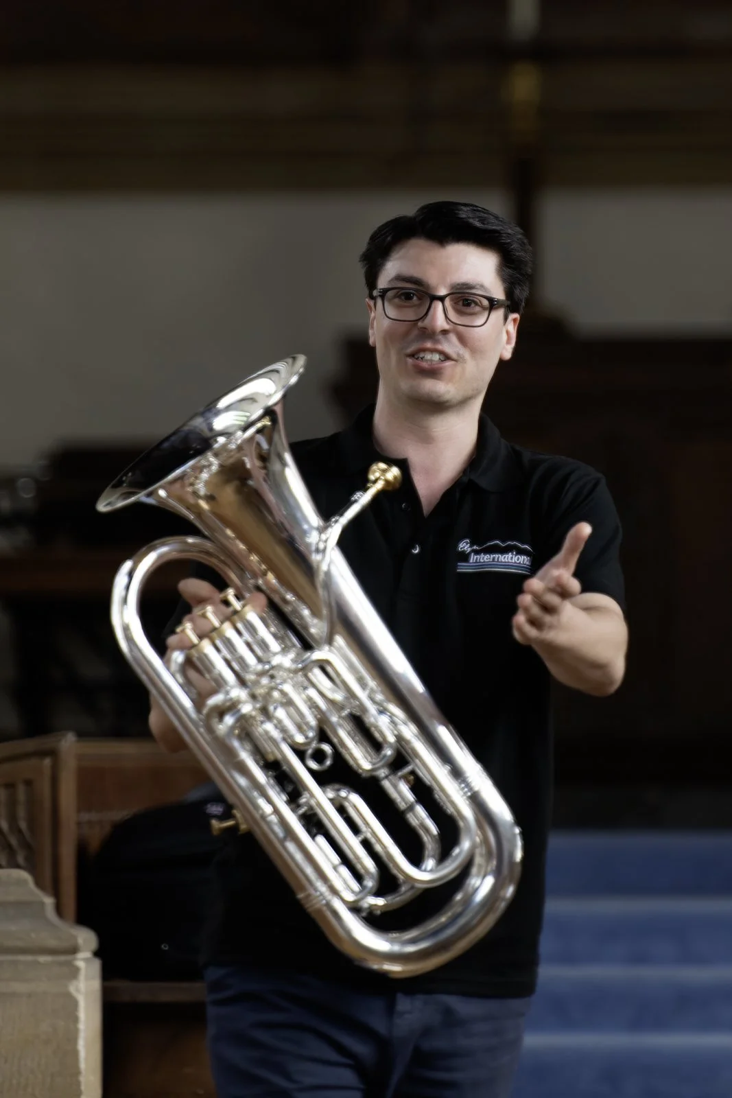 A man wearing glasses and a black shirt is holding a shiny brass euphonium and pointing with his index finger.