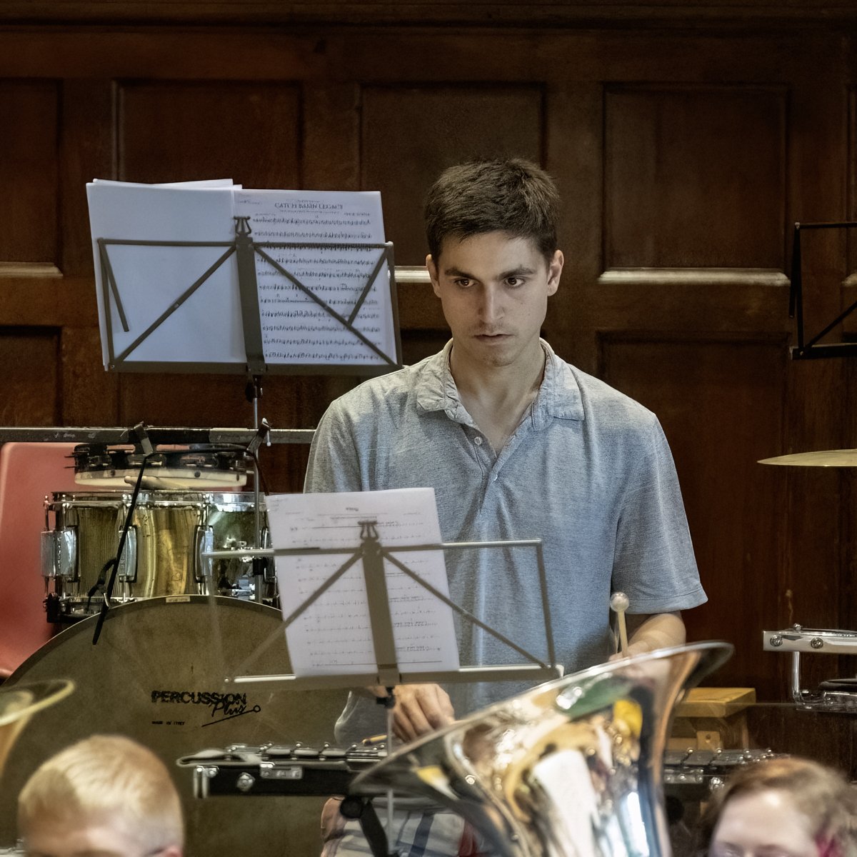A young man standing behind percussion instruments with sheet music on music stands in a wooden-paneled room.