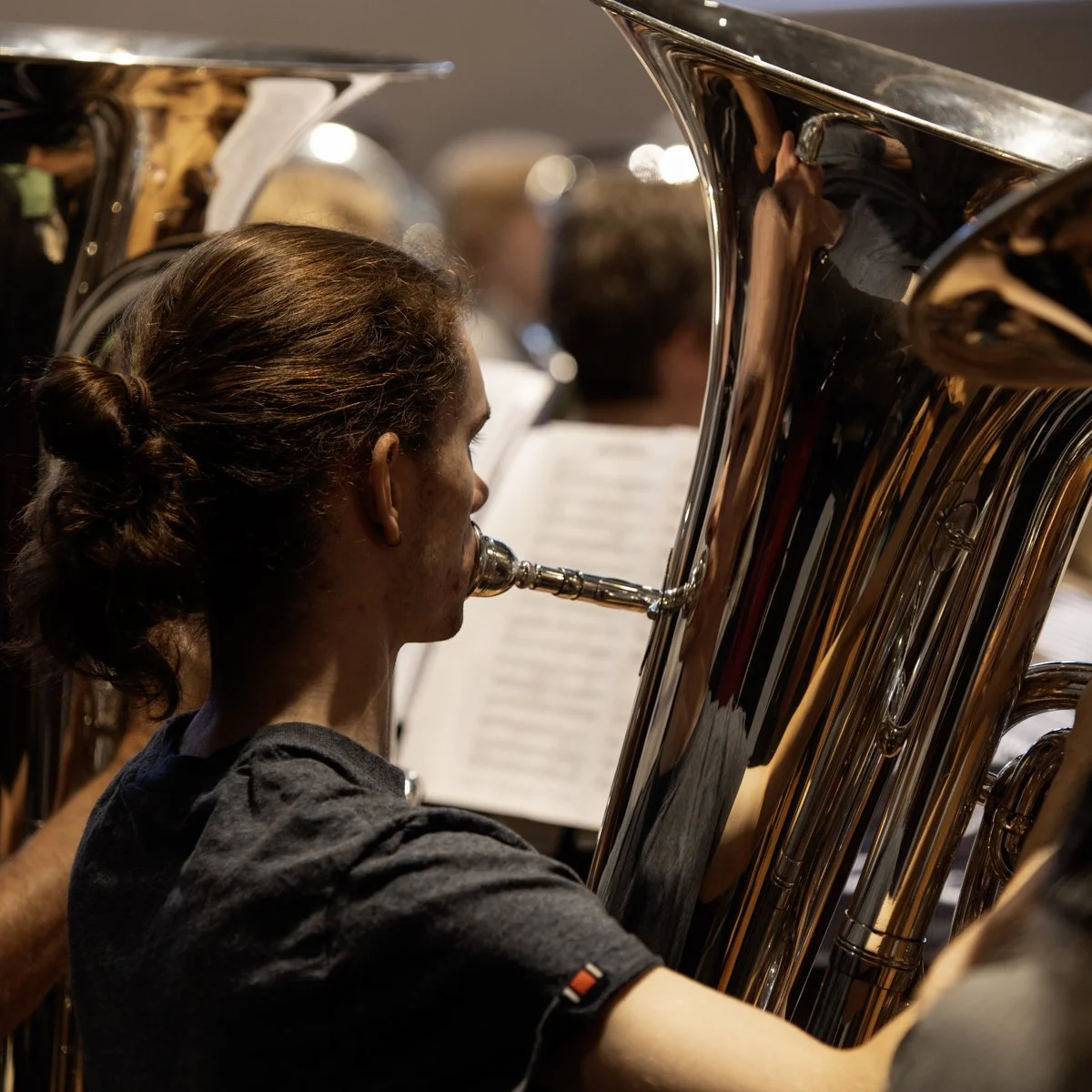 A person with long brown hair tied back is playing a brass instrument during a rehearsal, with sheet music in front of them.