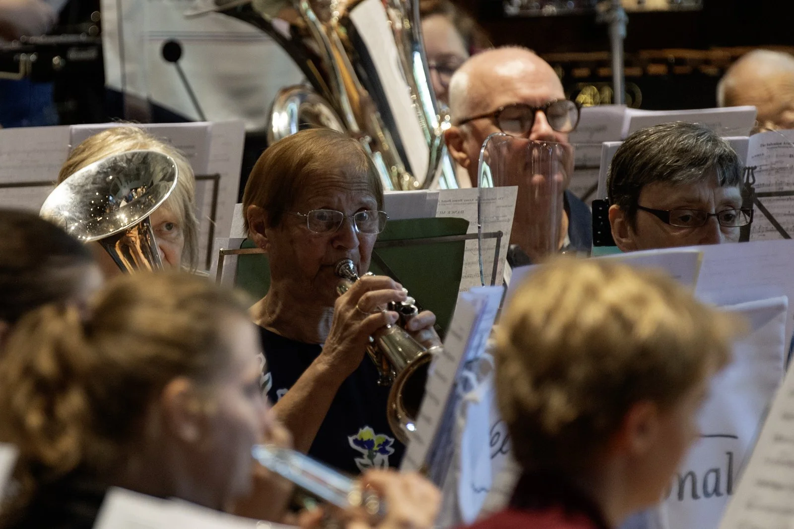 An orchestra performs with musicians playing flutes, horns, and other instruments behind music stands. The focus is on a woman playing the flute among a group of older musicians.