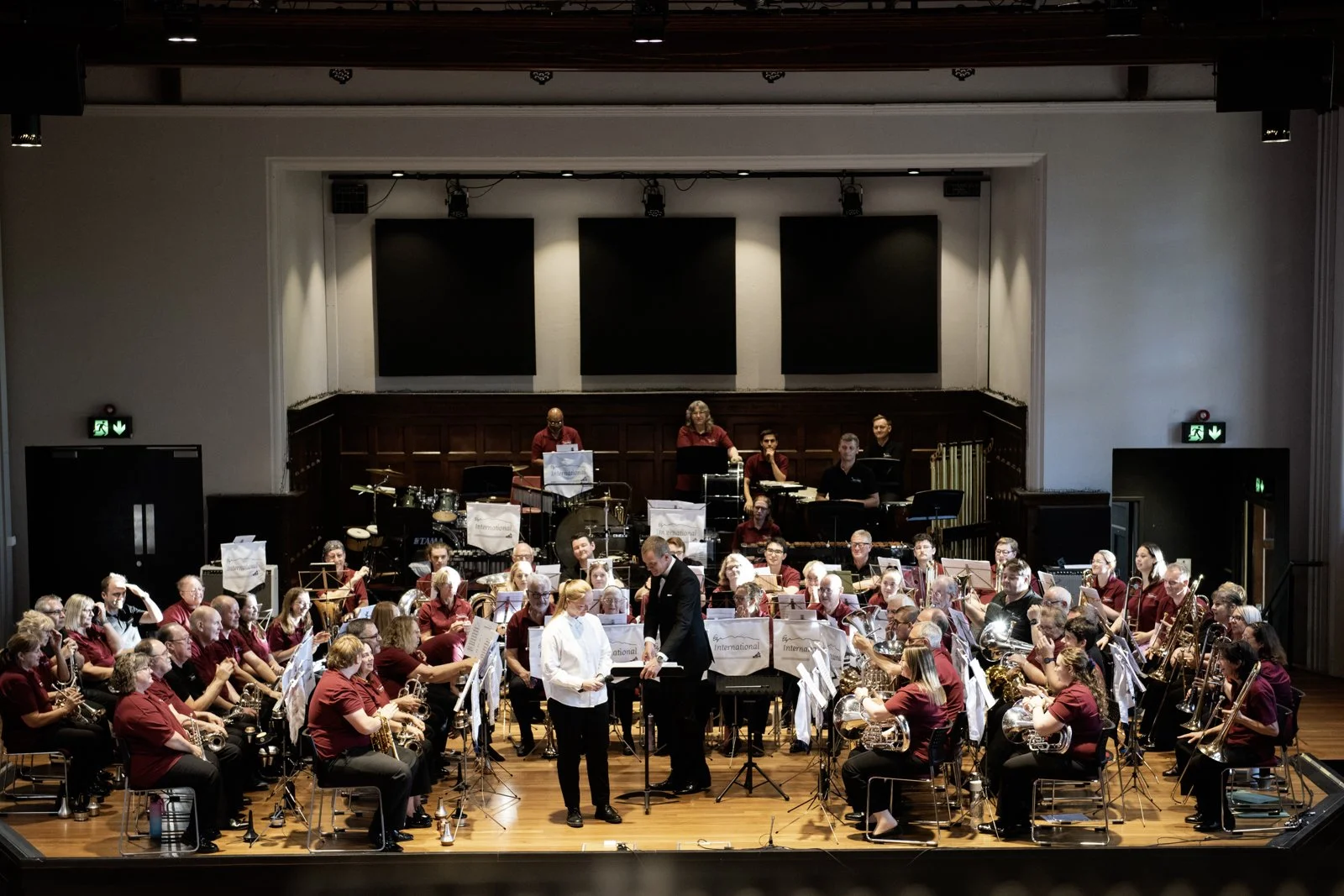 A large musical ensemble on stage performing with various instruments, a conductor and a soloist in front, in a concert hall.