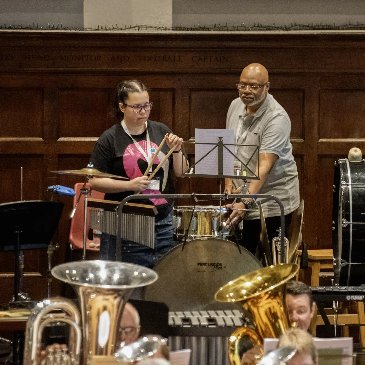 Two musicians, a woman and a man, are preparing to perform with percussion instruments in a wooden-paneled room with musical equipment and brass instruments in the foreground.