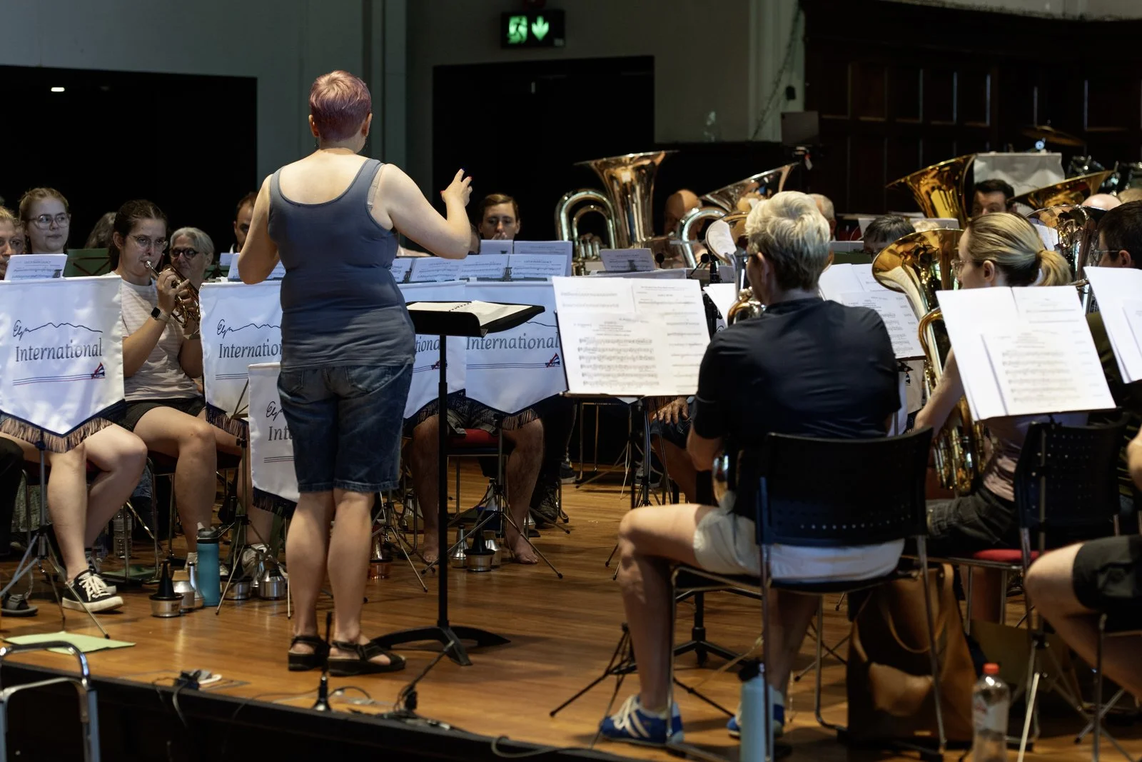 An orchestra rehearsal with musicians playing instruments, a female conductor standing, and music stands with sheet music on a stage.