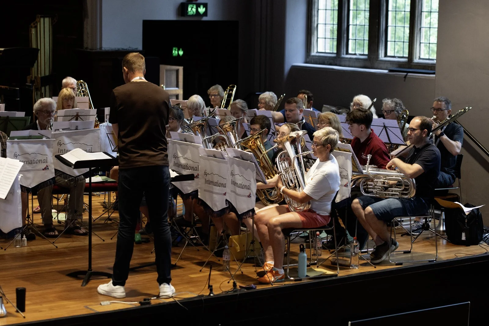 An orchestra rehearsing in a concert hall, with musicians playing brass instruments, a conductor standing in front, and large windows illuminating the scene.
