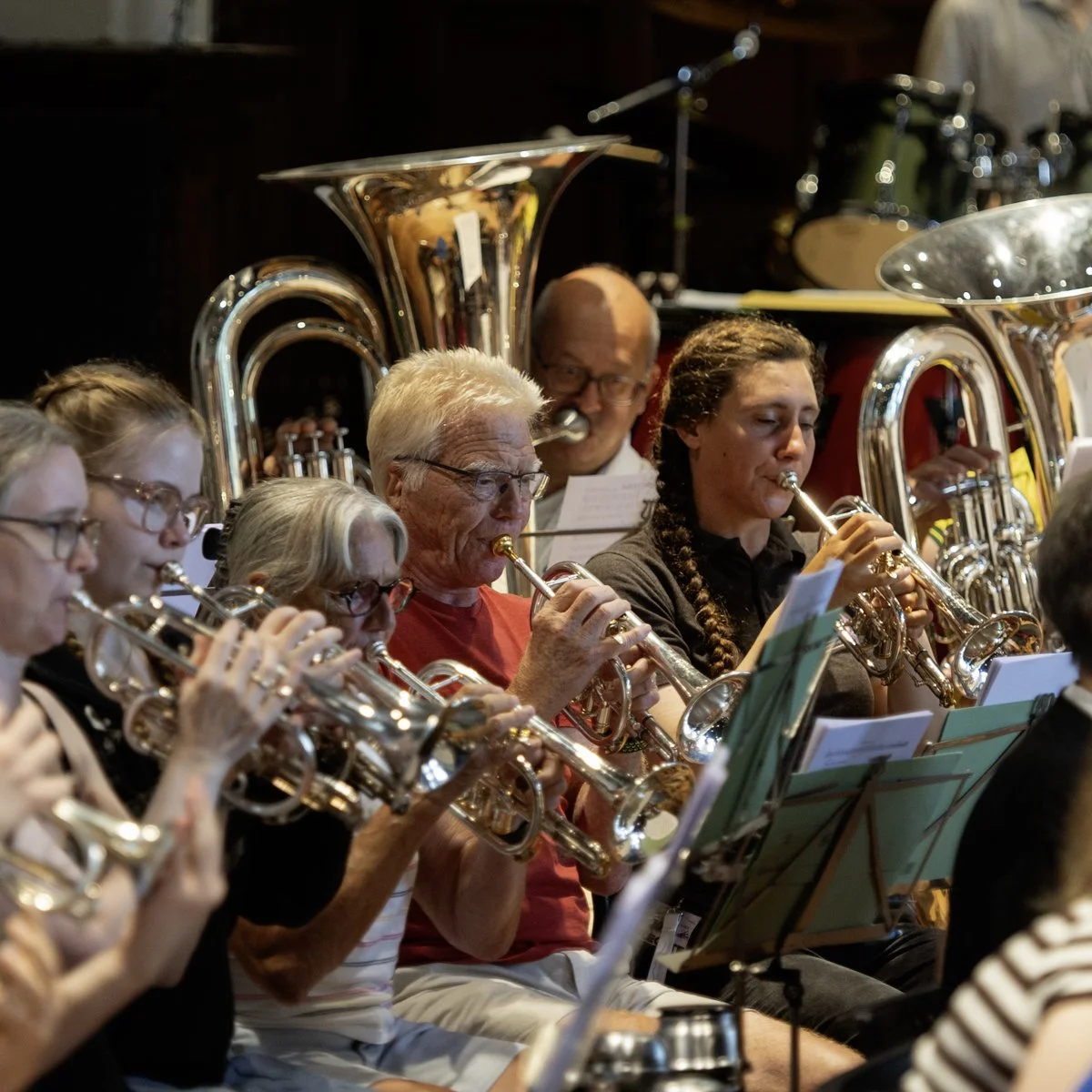 A group of musicians playing wind instruments, including trumpets and tubas, during a band rehearsal or performance.