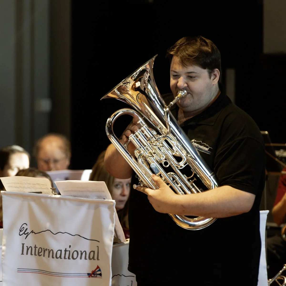 A man playing a silver euphonium during a concert, with sheet music and other musicians in the background.