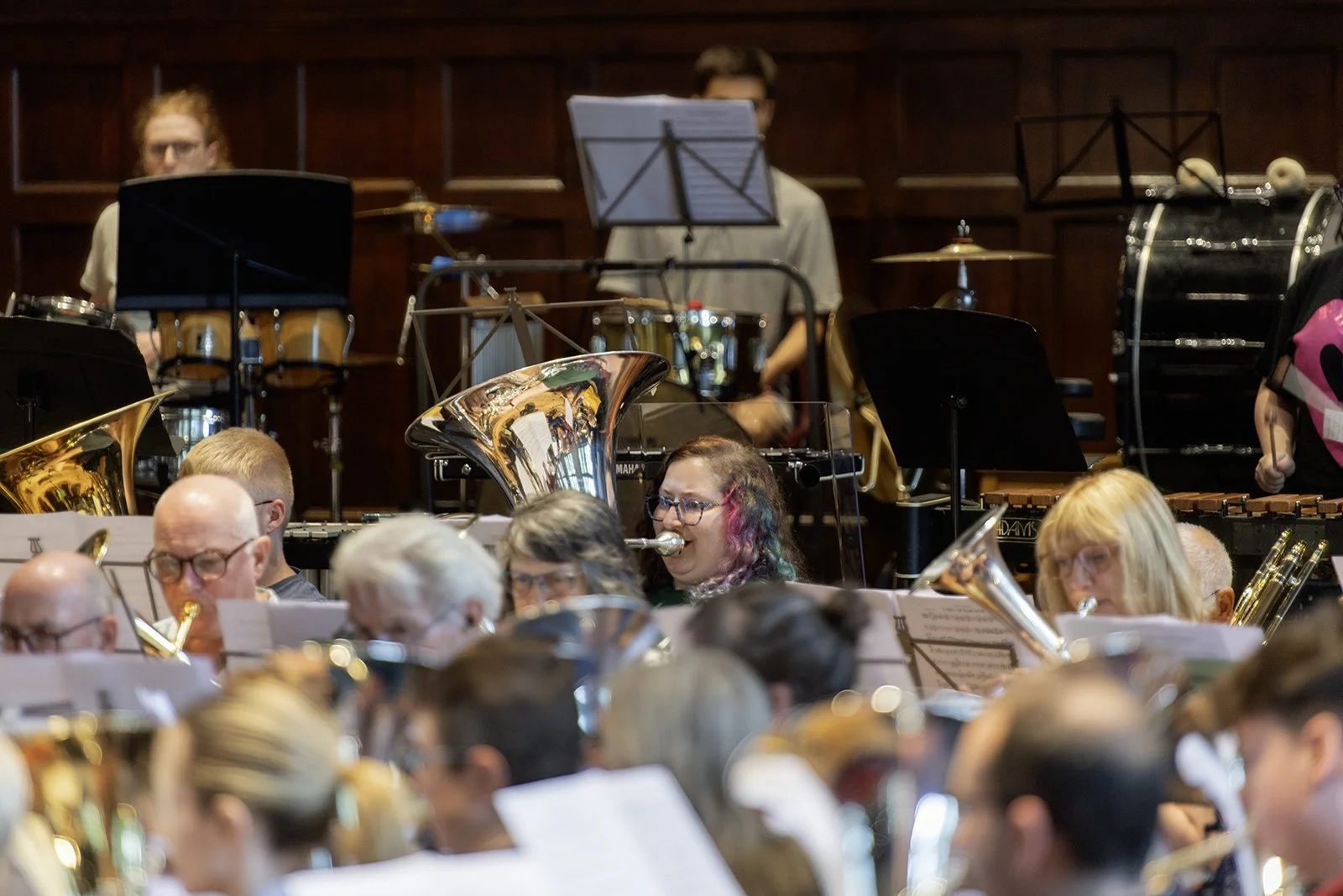 Symphony orchestra performing with musicians playing various instruments including tubas, drums, and xylophone in a concert hall with wooden paneling in the background.