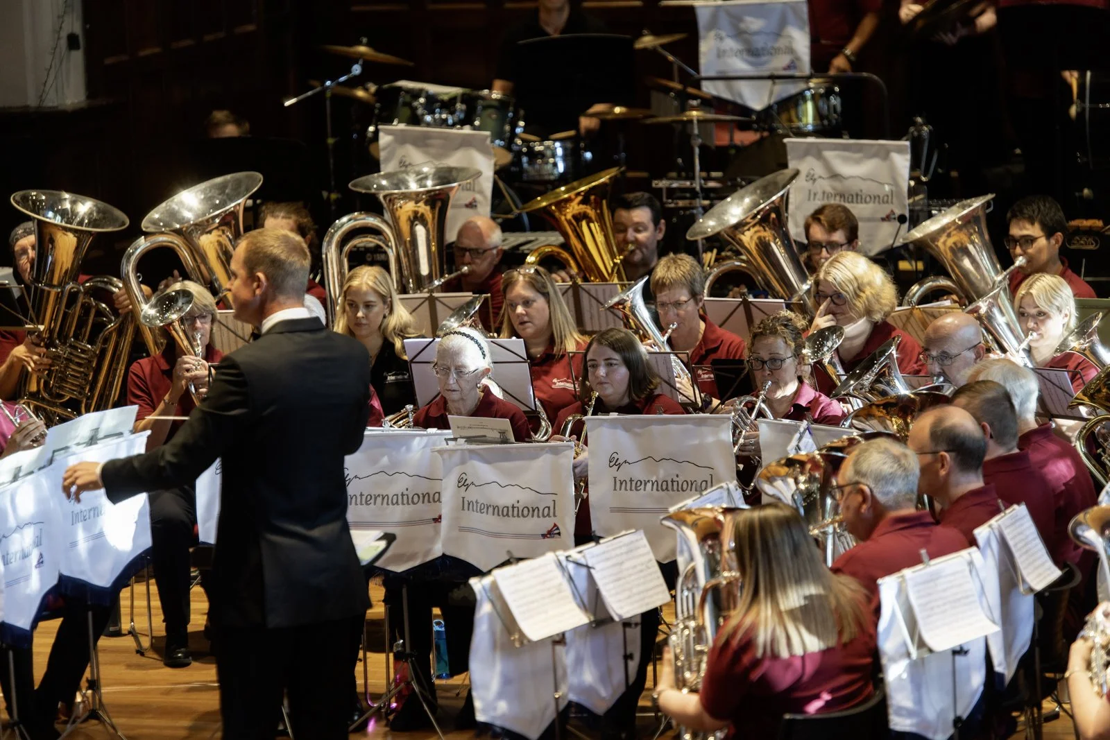 An orchestra performing on stage with a conductor in front, wearing a black suit and white shirt, leading the musicians who are playing brass, woodwind instruments, and drums. The musicians are dressed uniformly in red shirts and have music stands wi