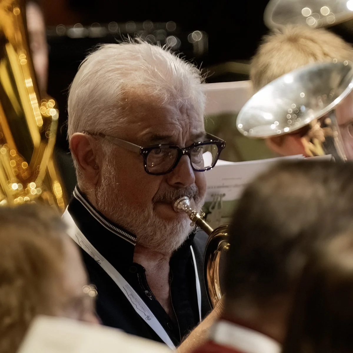 An elderly man with white hair and glasses playing the trumpet, surrounded by other musicians and music stands, during an indoor concert.