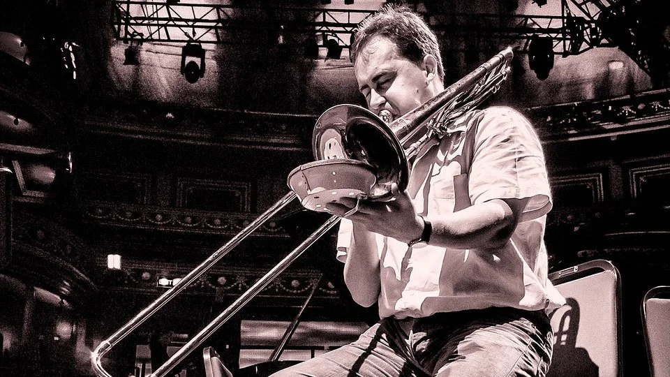 A man playing a trombone on stage, with a large theater or concert hall background, captured in black and white.