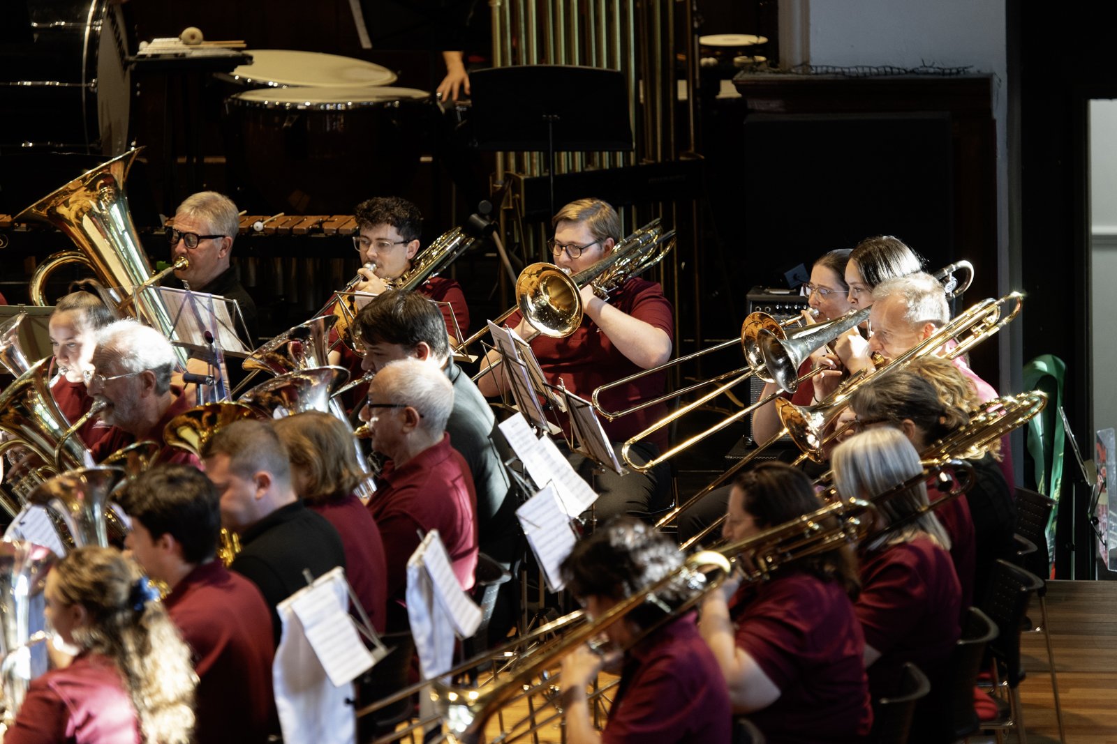 Orchestra performing in a concert hall, musicians playing brass instruments including trombones and euphoniums, with music stands and sheet music in front of them, dressed in red and black attire.
