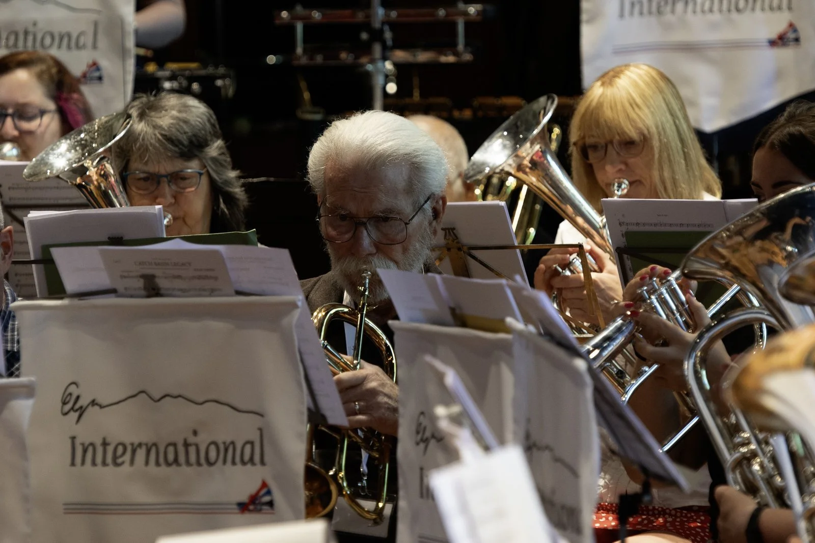 Group of musicians in an orchestra reading sheet music, with some playing brass instruments like tubas and trumpets; at the front, sheet music labeled 'International' is visible.