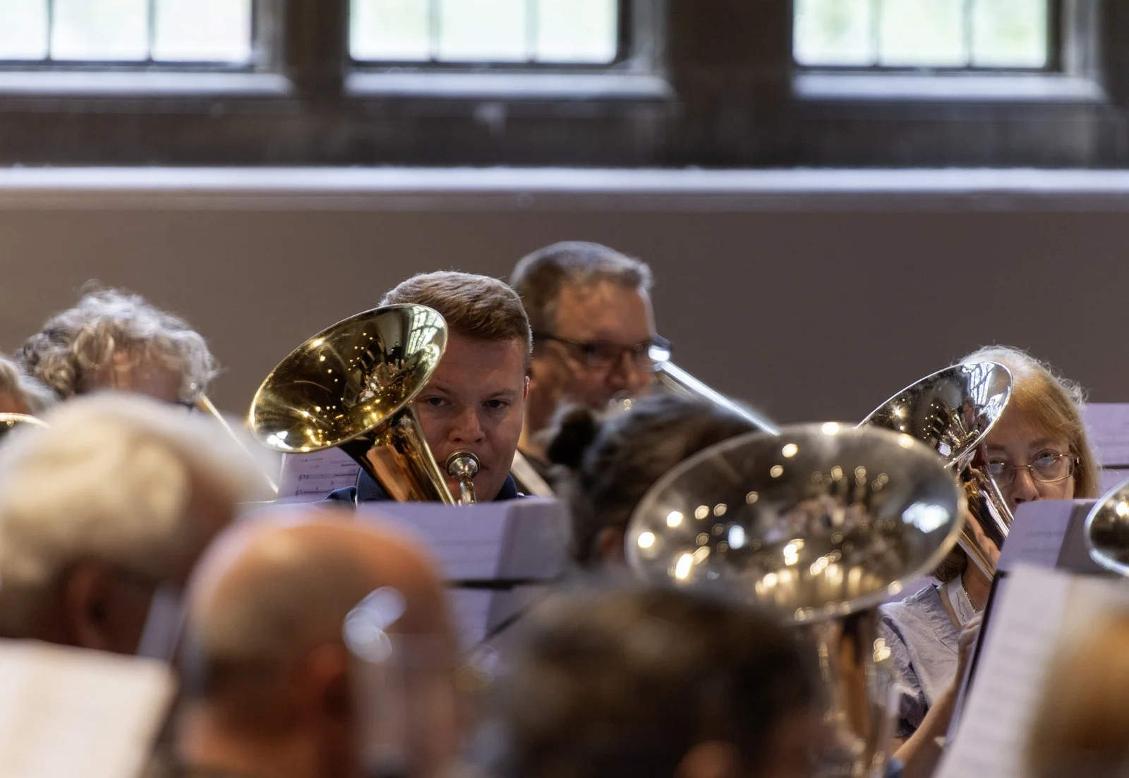 Members of an orchestra playing brass instruments during a performance inside a room with large windows.