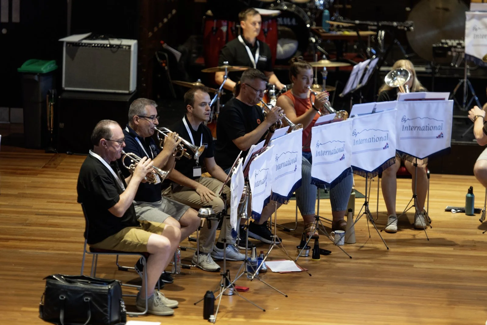 Orchestra members playing brass instruments during a concert in a hall with wooden flooring.