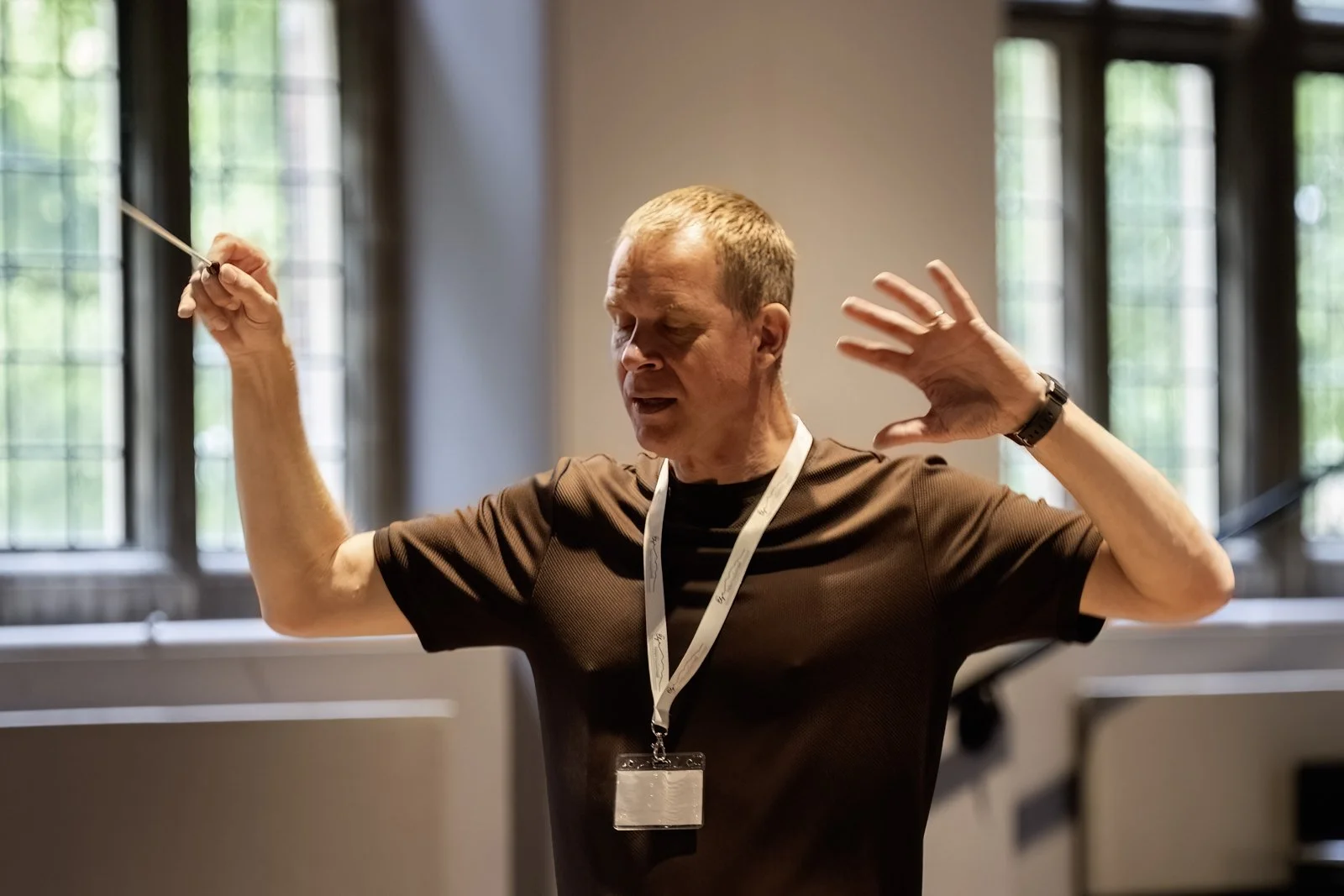 A man in a brown shirt with a lanyard around his neck, holding a pointer in one hand, raising his other hand near his face, speaking or singing, in an indoor setting with large windows in the background.