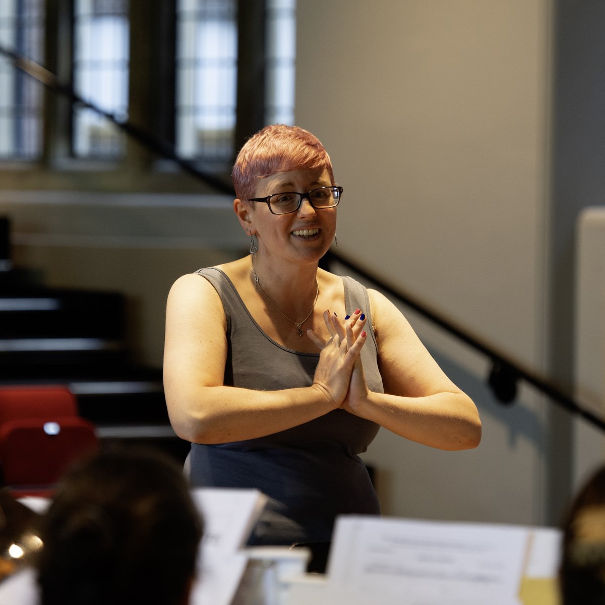A woman with short pink hair, glasses, and a gray sleeveless top smiling and clasping her hands together during a presentation or class, with people in the foreground and a staircase in the background.