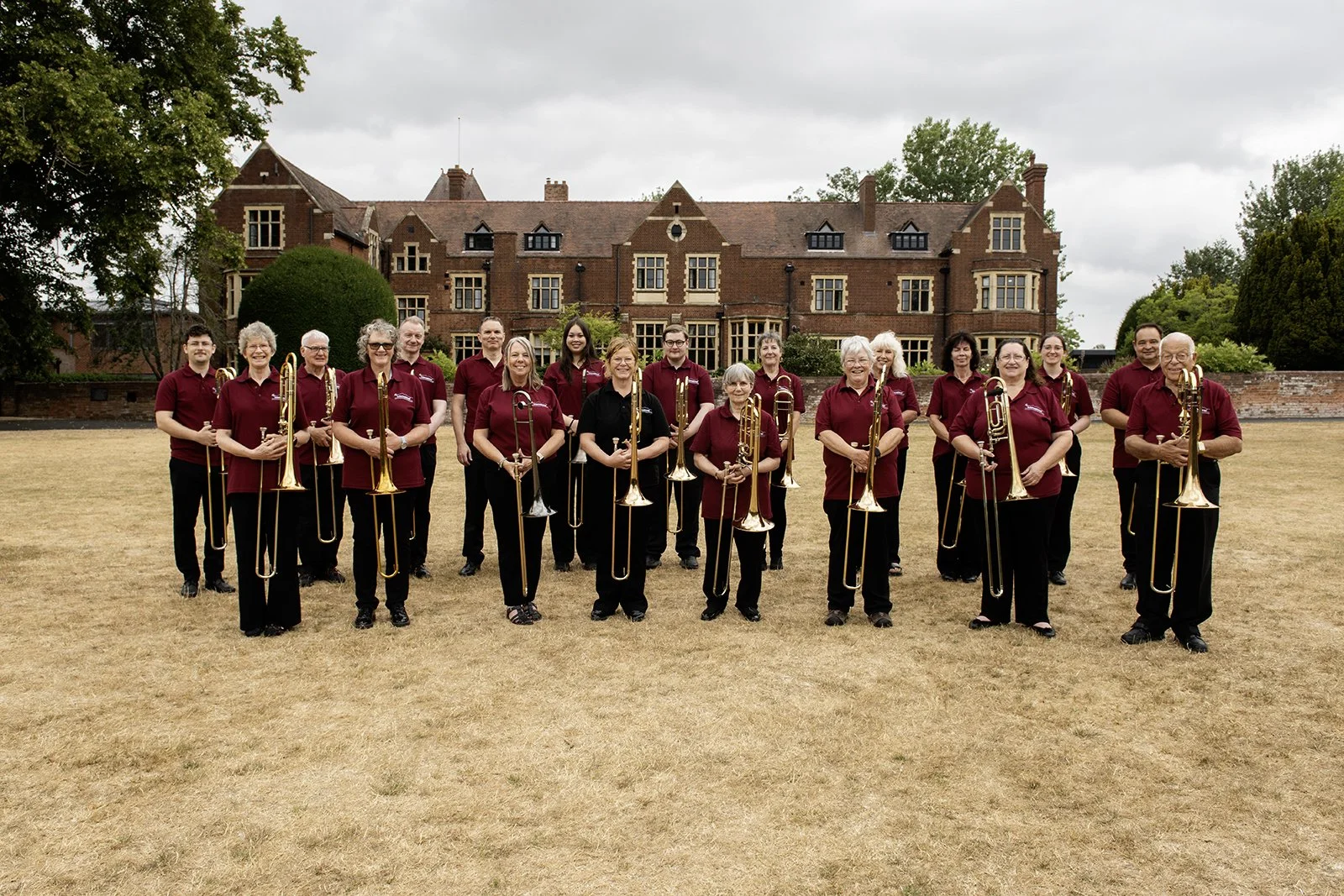 A group of people holding brass musical instruments, standing outdoors on a dry grass field, with a large historic brick building in the background.