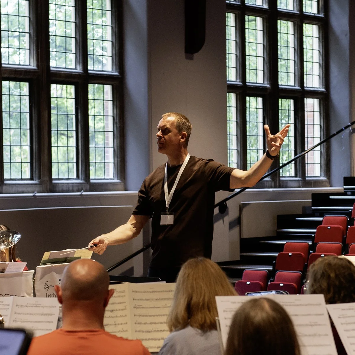 Man conducting an orchestra in a rehearsal space with large leaded glass windows.