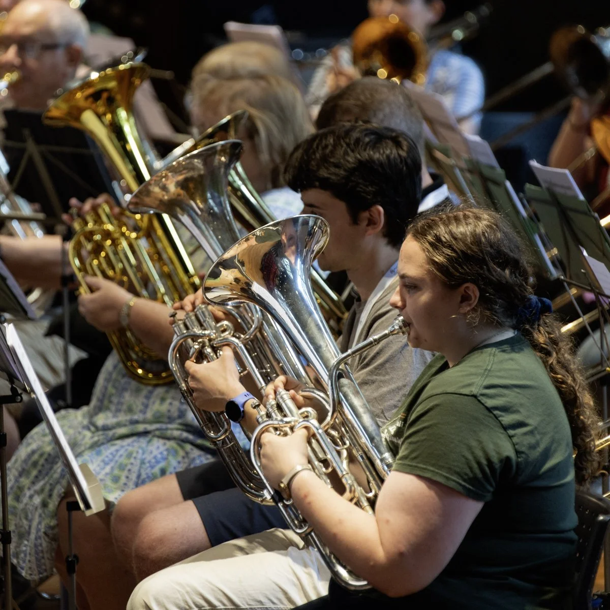 A group of musicians, including a young woman and young man, playing brass instruments such as euphoniums or tubas during an orchestra rehearsal or performance.