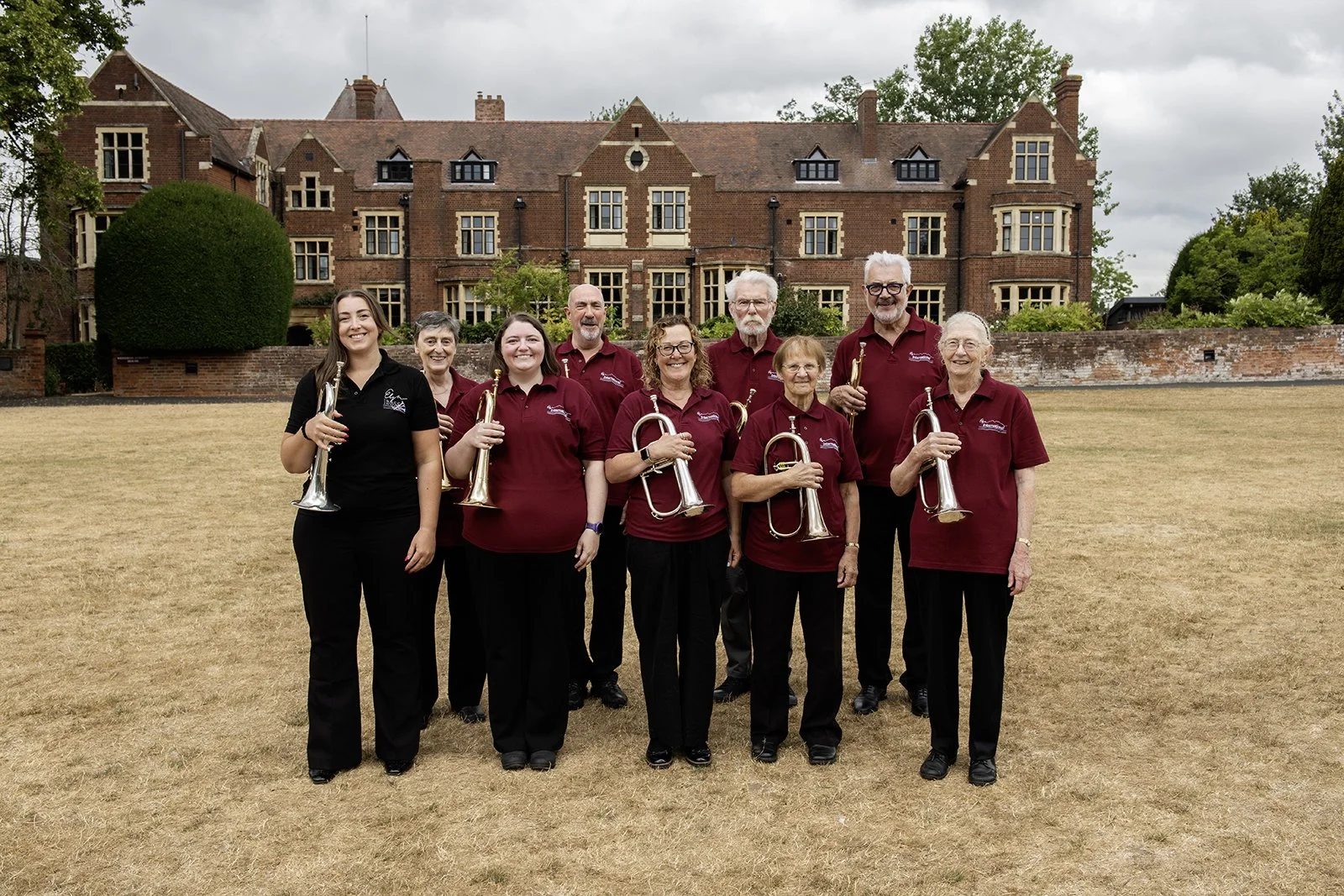 Group of nine people standing outdoors in front of a large brick building, holding brass musical instruments, smiling for the camera.